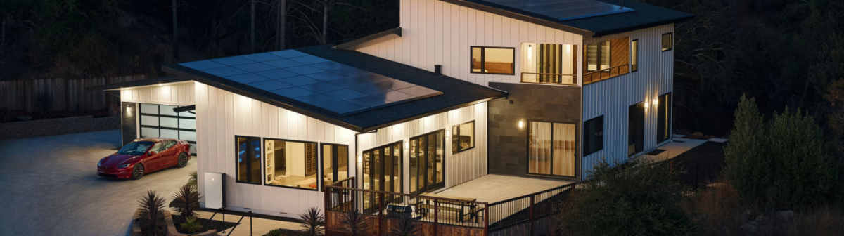 Modern two-story home with rooftop solar panels, large windows, and a red electric car parked in the driveway at dusk, surrounded by natural landscaping and trees.